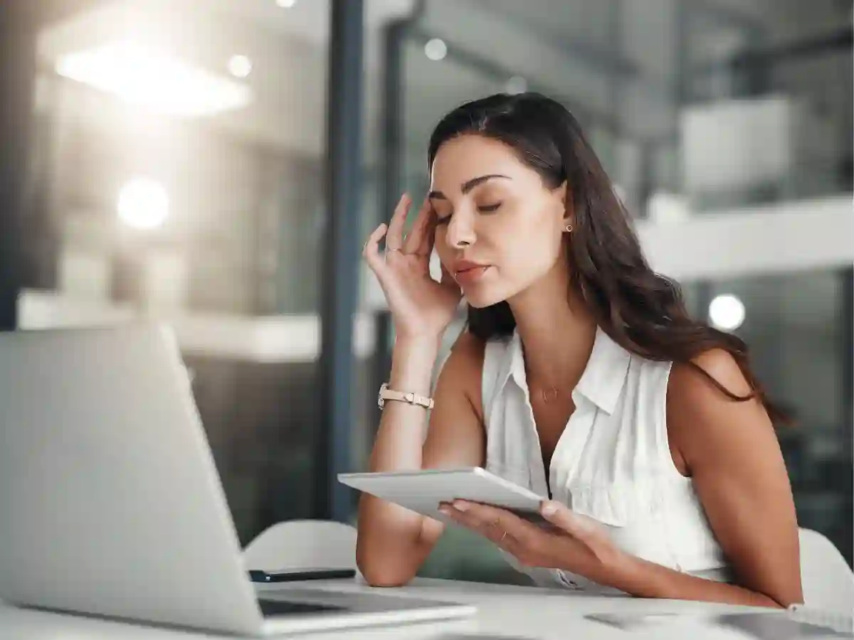 Woman sitting at a desk rubbing her temple with one hand while holding a tablet in her other hand