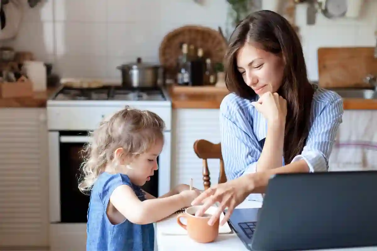 Mom working at home with laptop on kitchen table smiling while her daughter draws in a notebook next to her